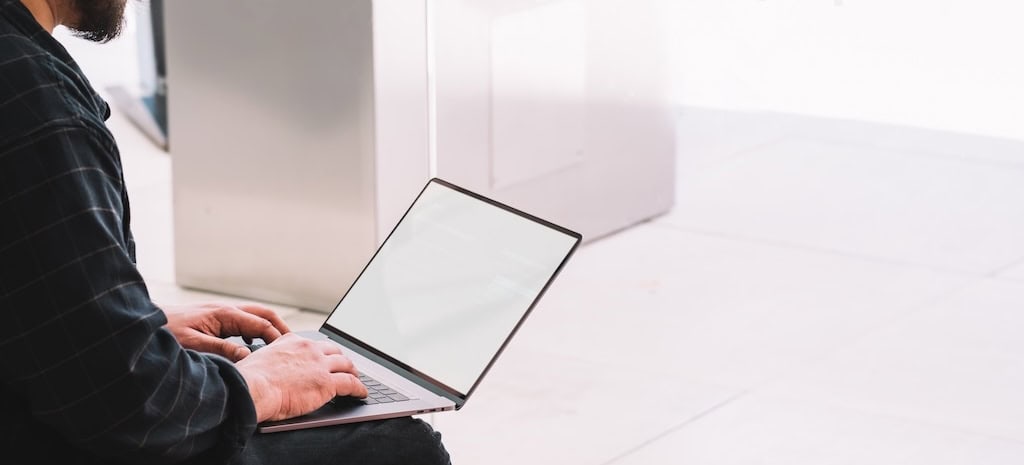 Side view of crop bearded male in casual outfit browsing laptop with empty screen while sitting on sofa in contemporary IOP program